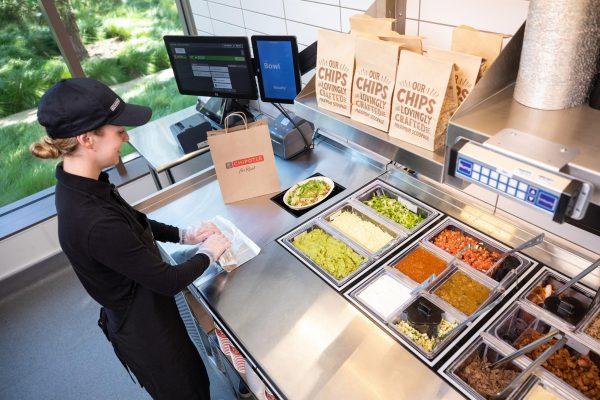 A Chipotle team member rolls a burrito while the automated makeline assembles a bowl. Burritos, tacos, quesadillas and kids meals can be made above the makeline while the automated system creates bowls and salads. 
