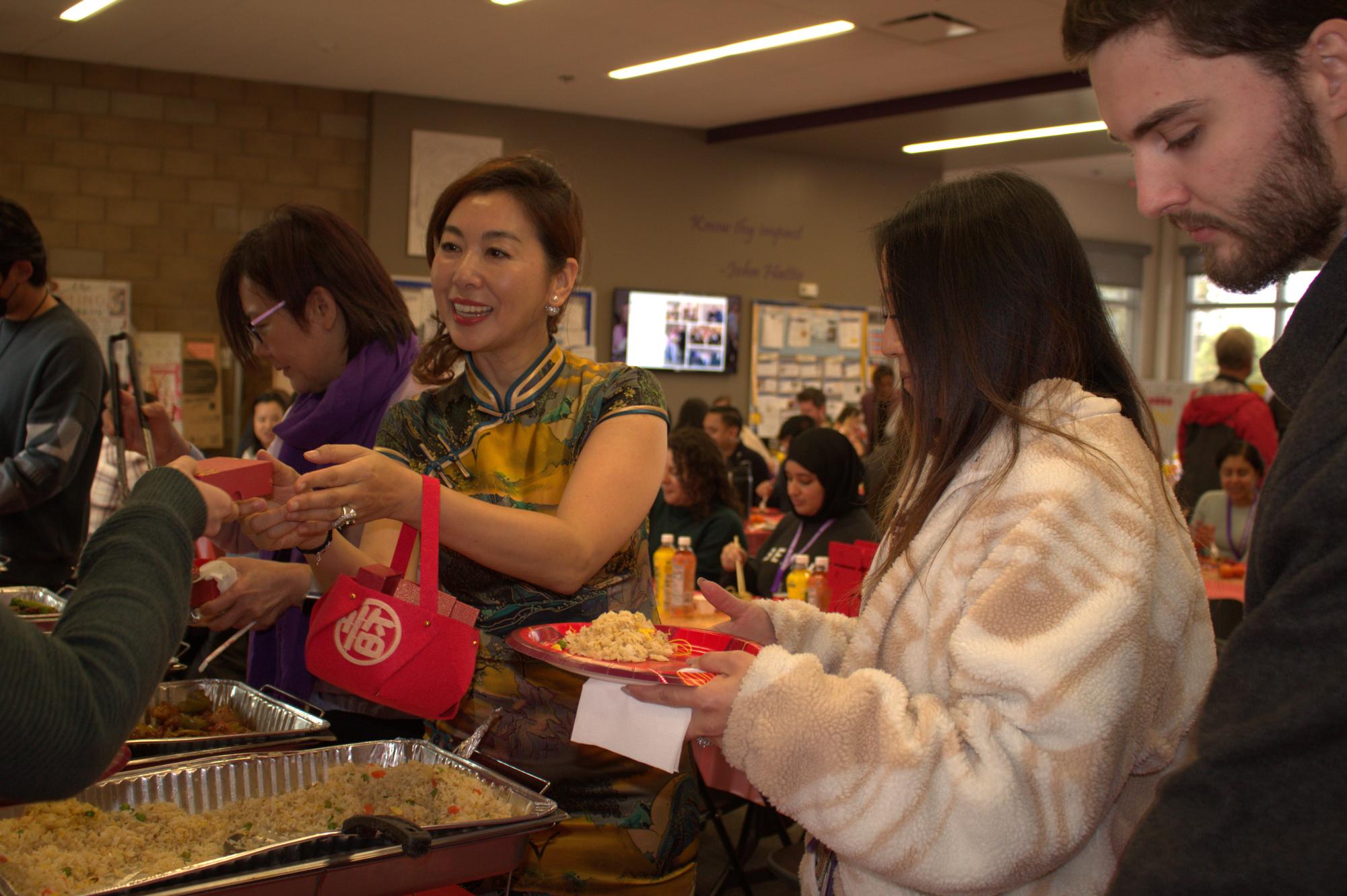 Chinese Parent Group member KK Chiang hands a staff member a custom USB holder featuring a Chinese tassel and an engraving of Buster the Bulldog in the staff lounge, where Chinese food is being served.
