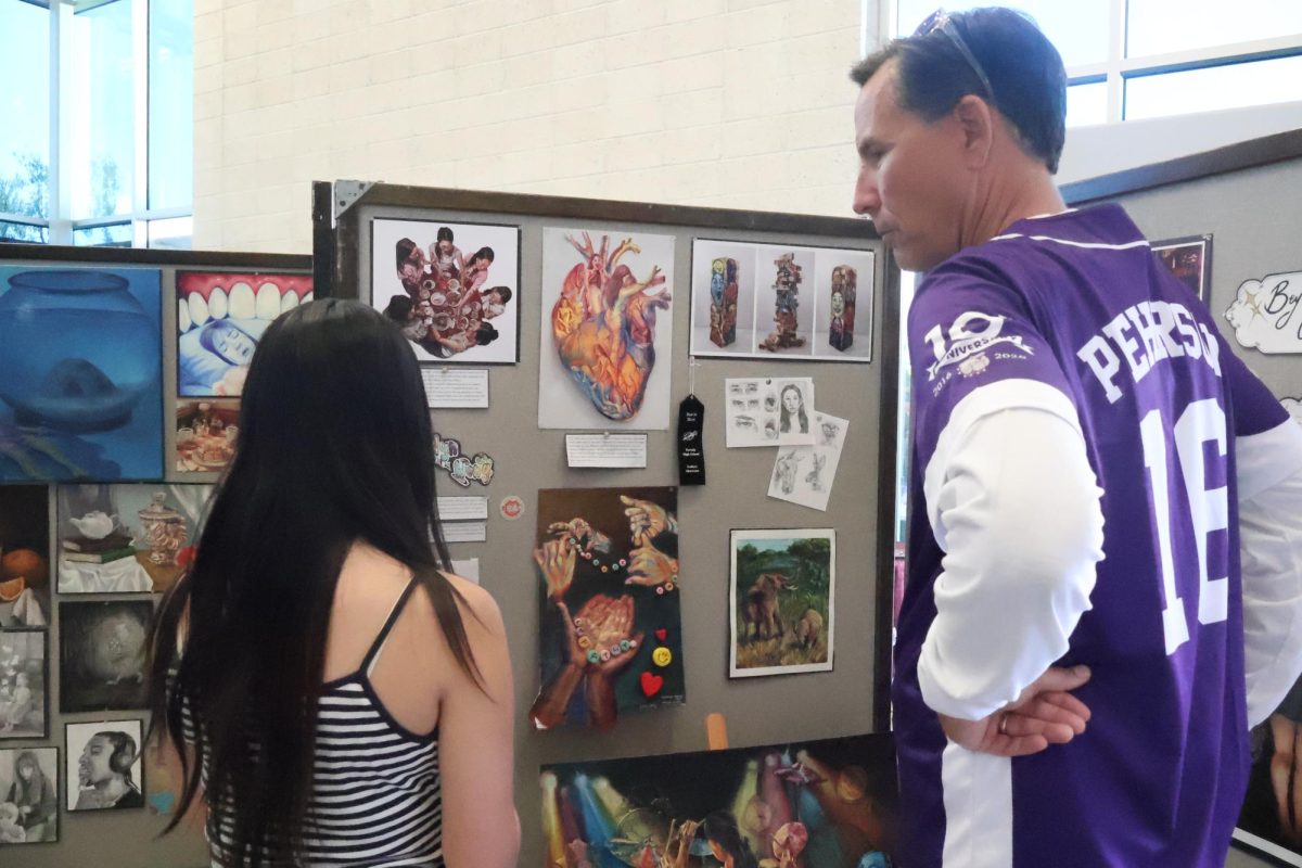 Portola High School's Visual Arts Program presents its annual showcase, Night of the Arts, on April 10. At the exhibition, artists display their creations from the school year on boards, creating thematic exhibitions with various media showcases. Junior Katelyn Wong and Principal John Pehrson observe her display, which combines interactive elements like Jenga with 2D artwork.